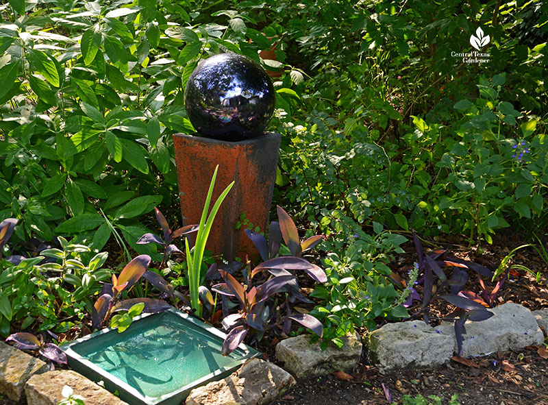 various plants in dappled shade with green platform water bowl, rust-colored planter and purple gazing ball 