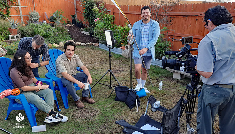 men and woman in backyard garden with TV camera, lights, and microphones
