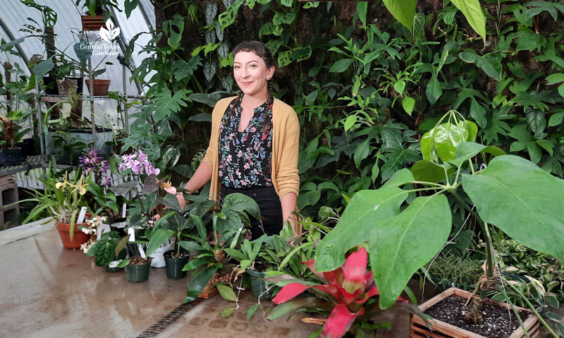 smiling woman in greenhouse with many plants on table in front of her