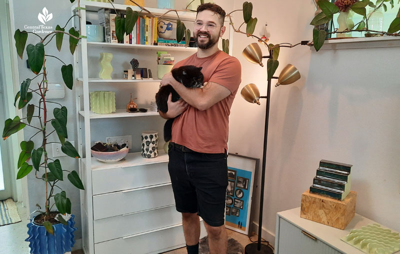 man holding cat in front of bookshelf holding various ceramics 