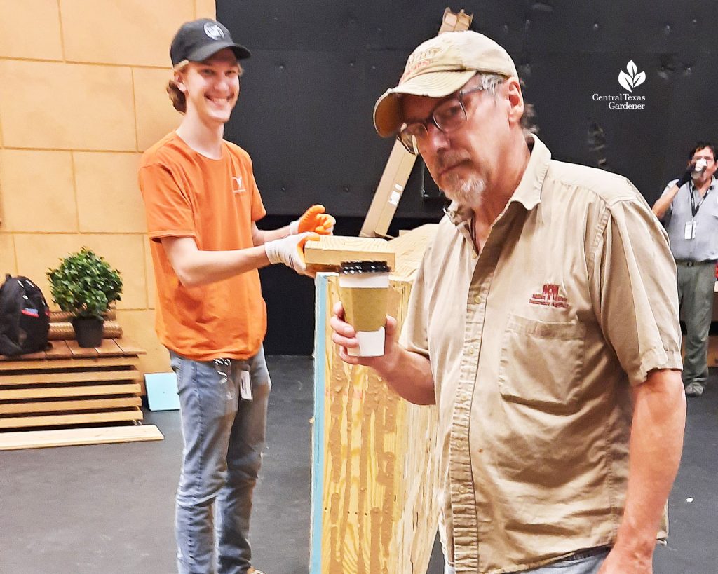 man with coffee cup smiling at camera; college student working on set behind