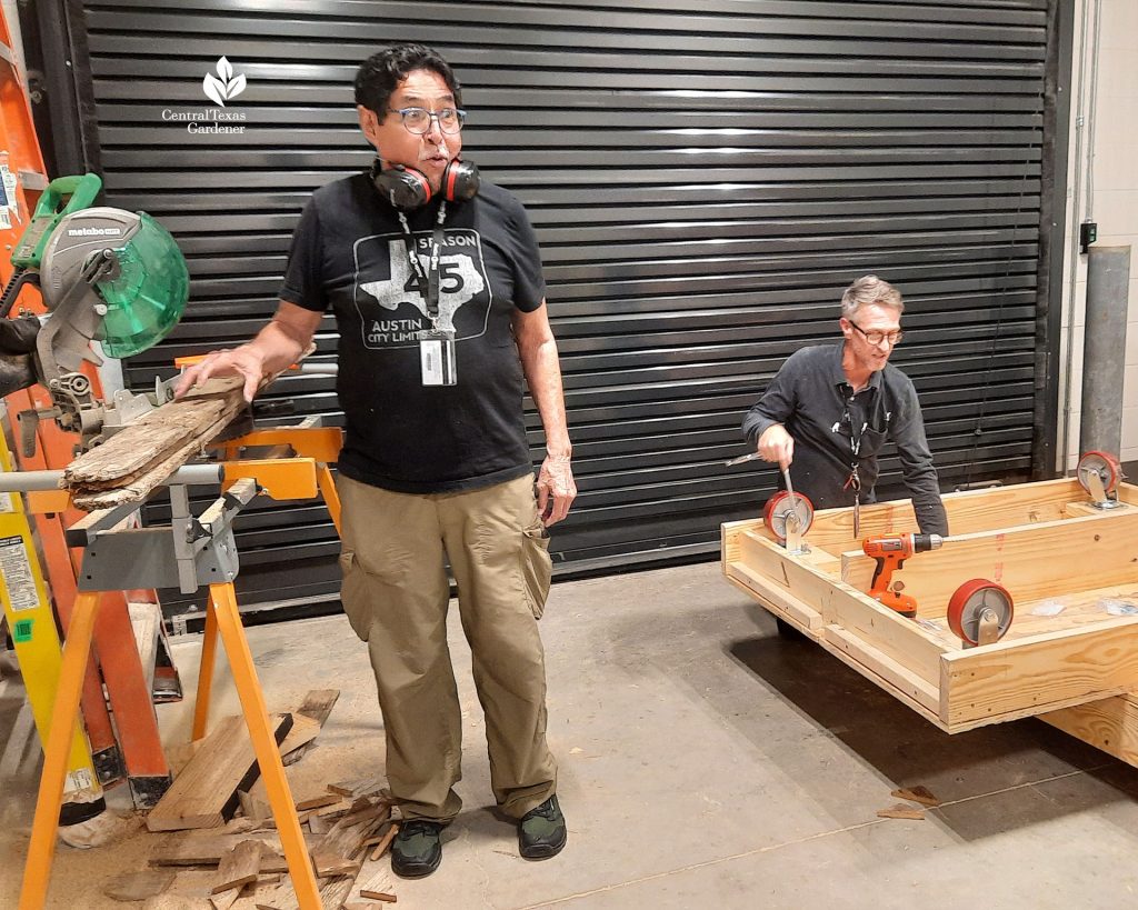 one guy with headphones around neck standing next to electric saw and another man adding casters to platform