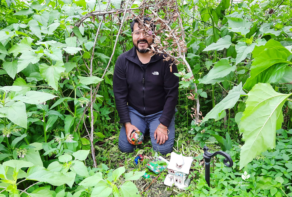 man smiling in kid hut of broken branches; toys on ground, sunflowers surround him