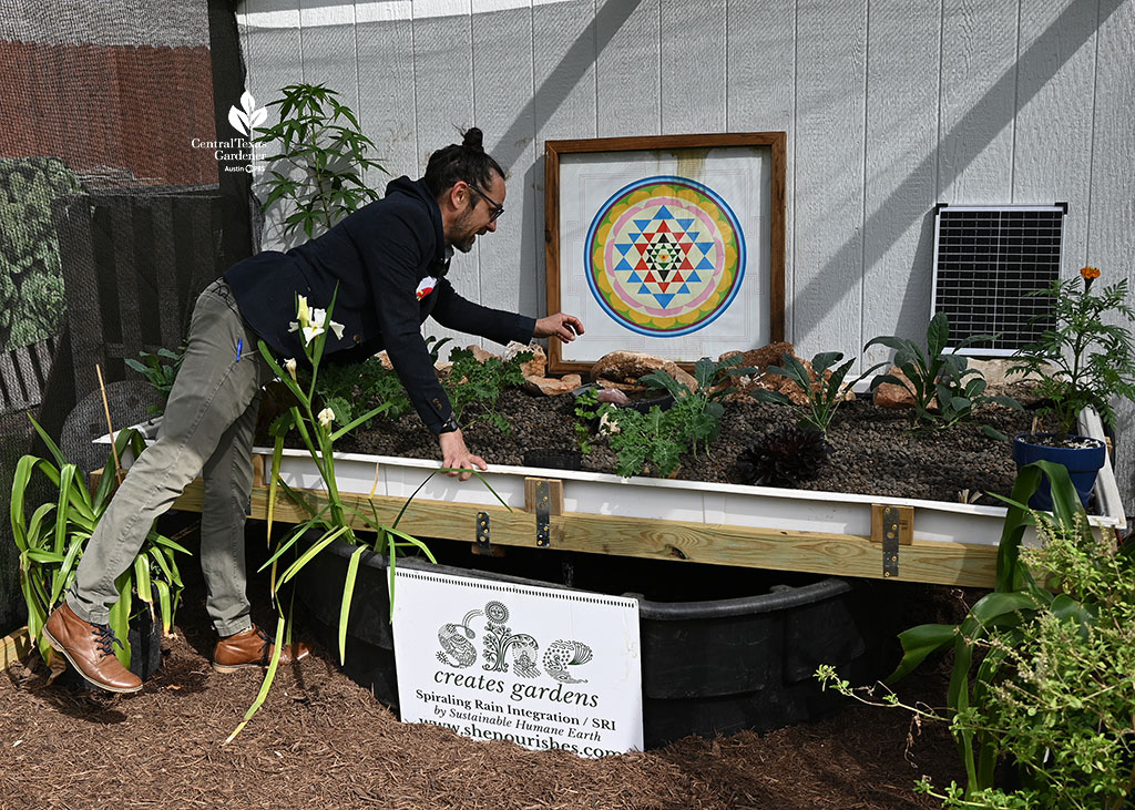 man leaning over waist-high aquaponics bed with SHE sign in front for Sustainable Humane Earth
