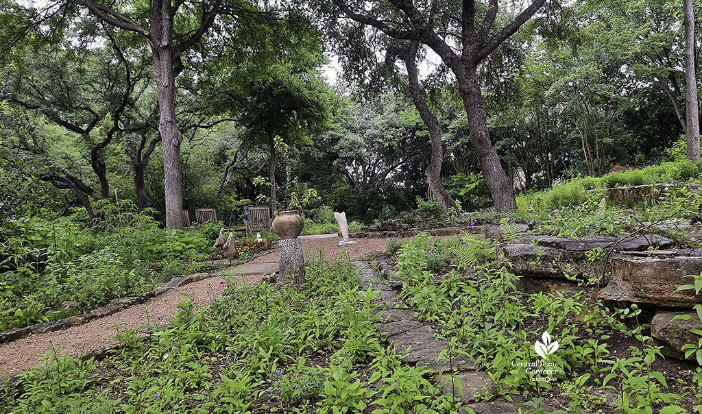 gravel path and stone path bordered by plants lead to a pedestal-mounted limestone that resembles a dolphin 