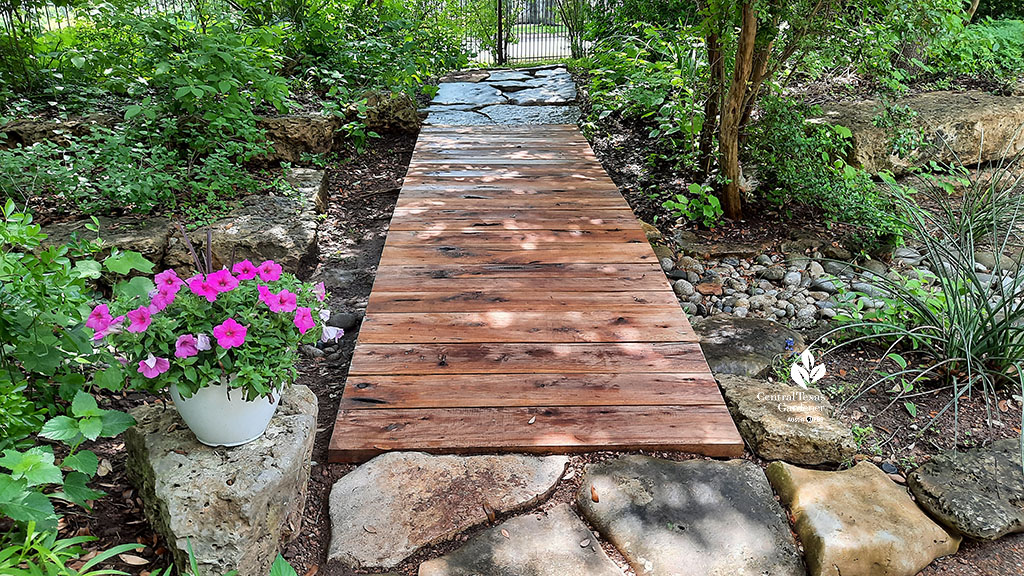 multi-toned wooden bridge over dry creek bed framed by native plants and flowers in containers 