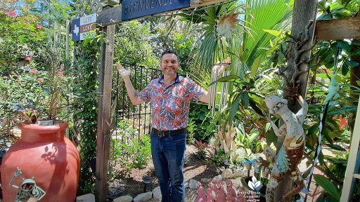 man standing in backyard garden entrance with sign that reads Bienvinedo