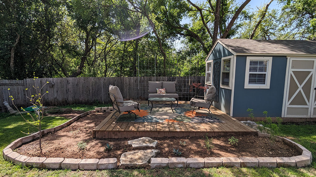 new planting bed around a shed and small raised patio deck. A colorful rug, a glider, and two chairs sit on the deck. 