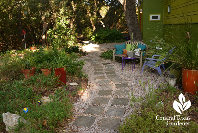 flagstone path bordered by plants and bench and chair with colorful cushions. 