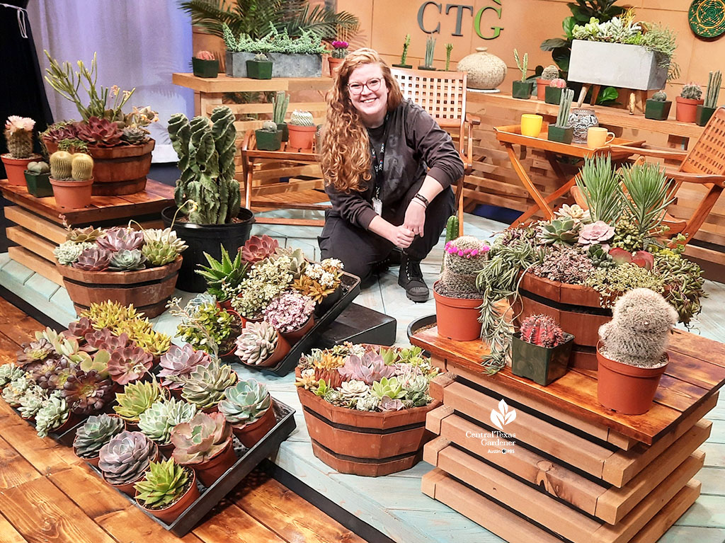 smiling young woman kneeling on CTG set with flats and containers of multi-colored succulents and cactus