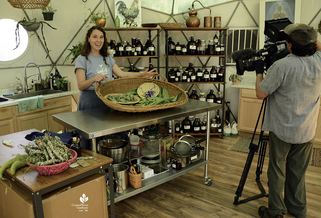 woman smiling at camera in room with large basket of herbs on a table and shelves of bottles behind her 