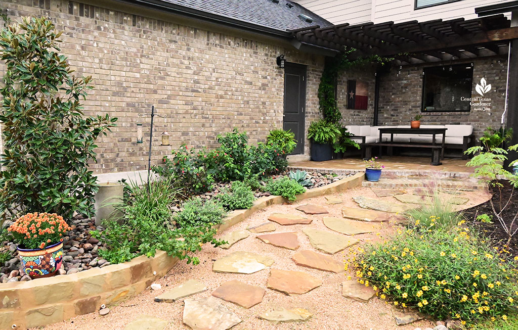 stone-lined garden bed next to house filled with young plants. A pathway of flagstones set on crushed granite ends at a wide patio 
