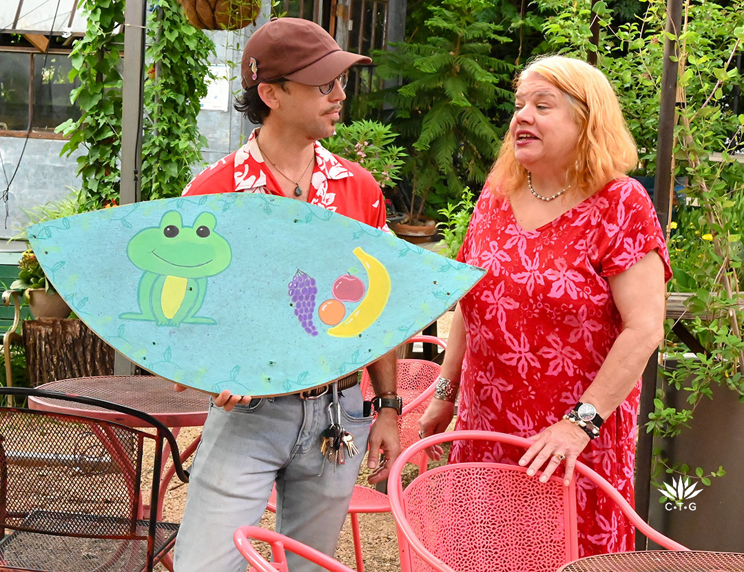 Young man holding brightly painted wood of a frog and fruit; showing it to 
Bonnie Cullum