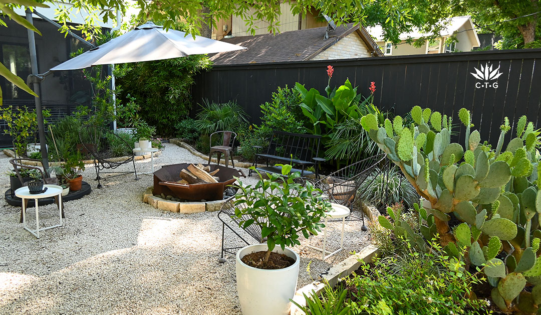 metal fire pit on gravel patio; bench against fence border of tall plants and blooming canna